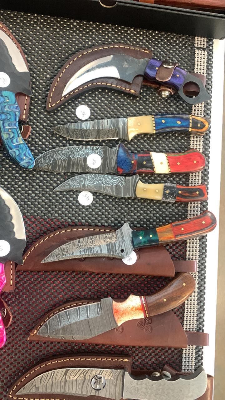 Display of handmade knives in leather sheaths, with colorful handles and curved blades.