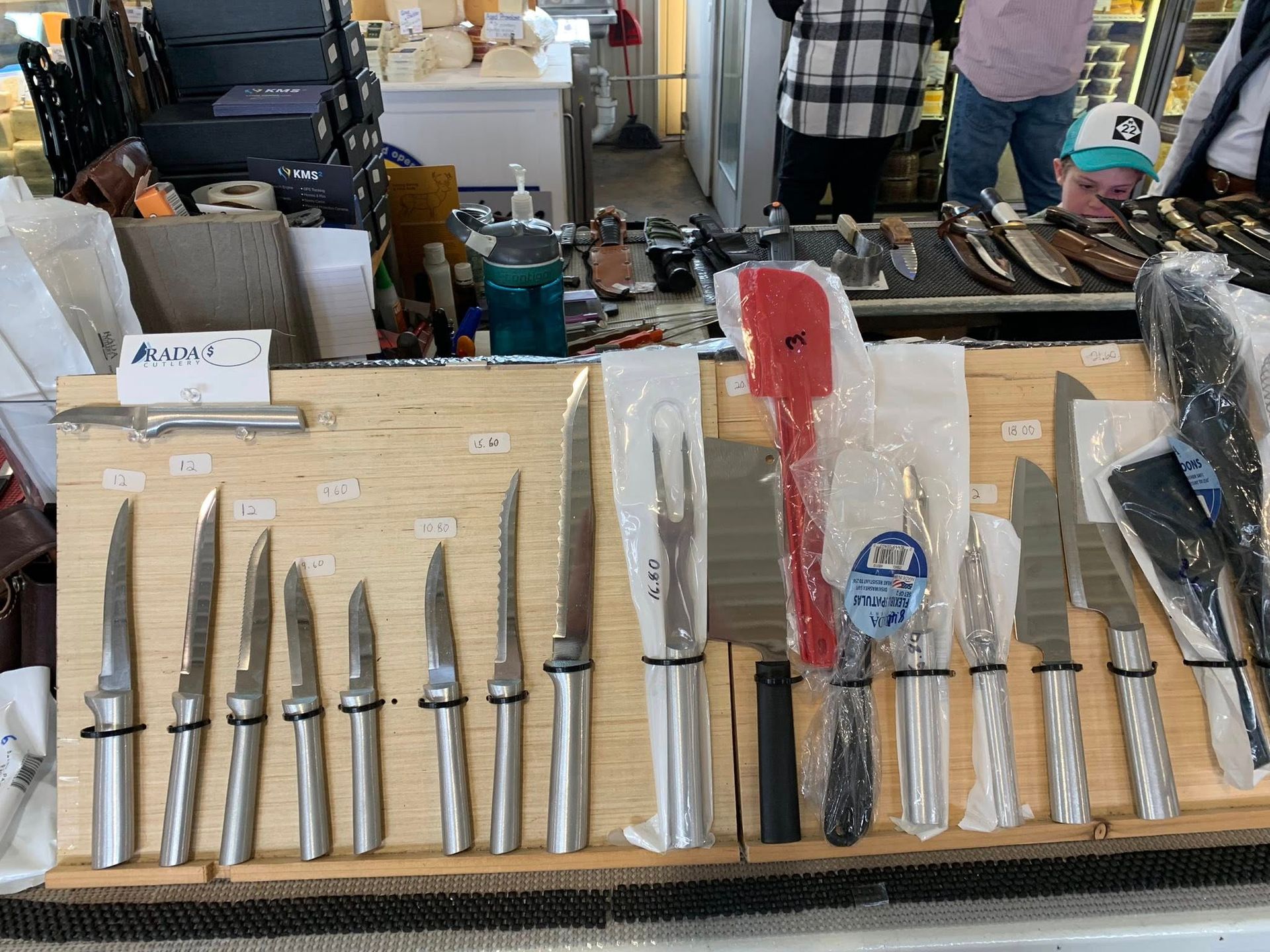 Knives displayed on a wooden surface at a market, with various shapes and sizes, plus a red spatula.