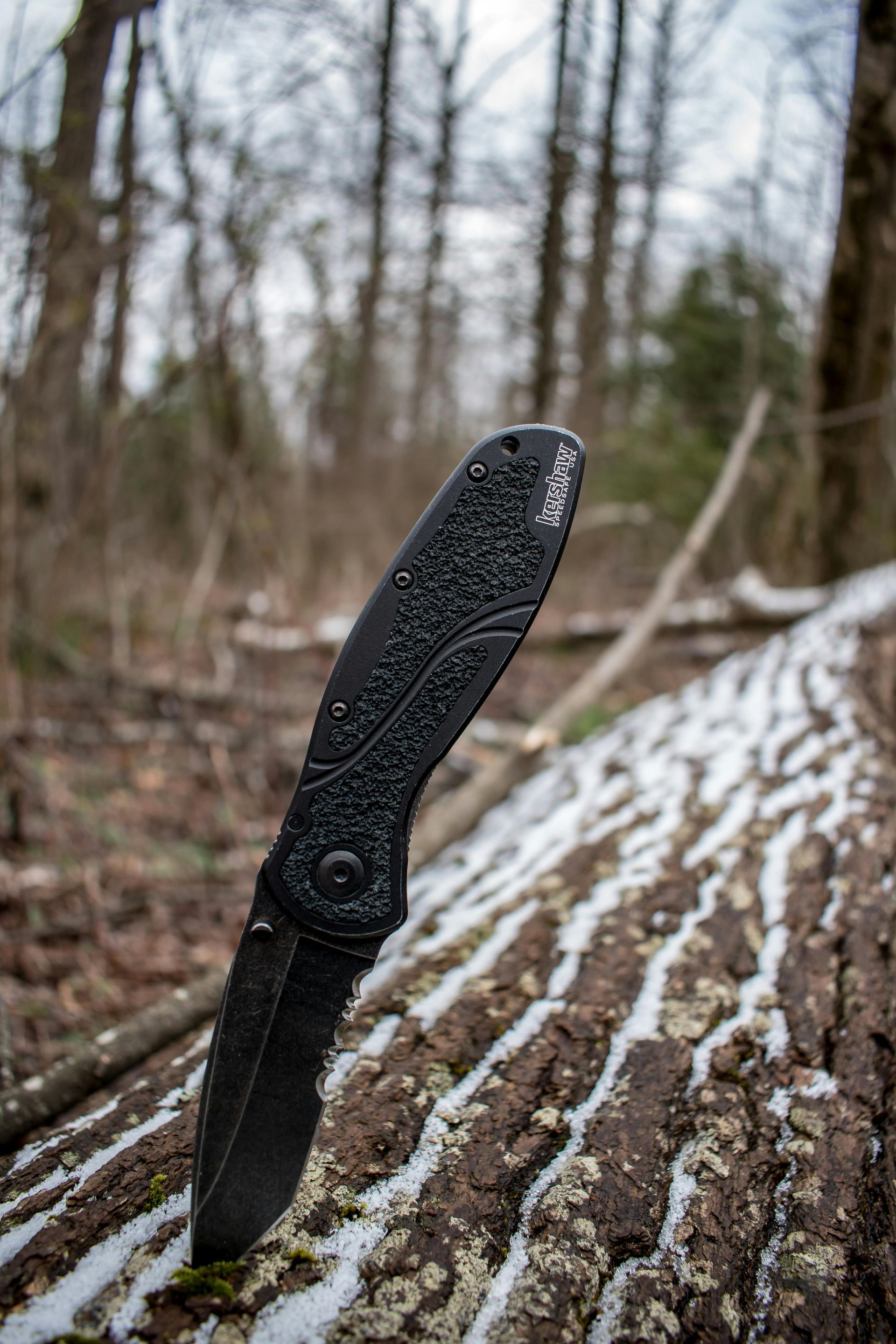 Black folding knife stuck in a snowy log in a forest setting.