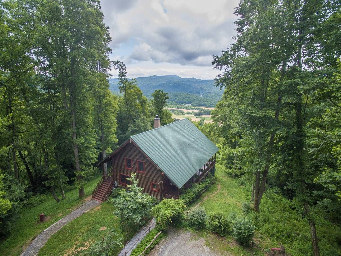 Wooden cabin with green roof nestled in trees, overlooking a mountain valley under a cloudy sky.