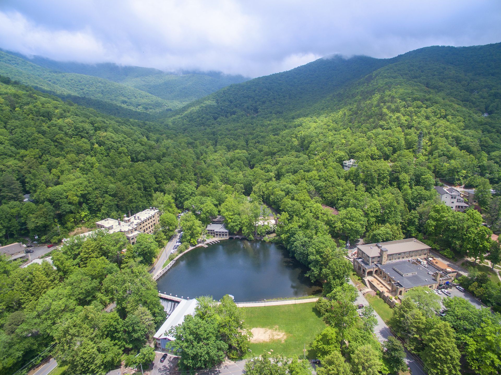 Aerial view of lake surrounded by green trees and mountains. Buildings sit at lake's edge.
