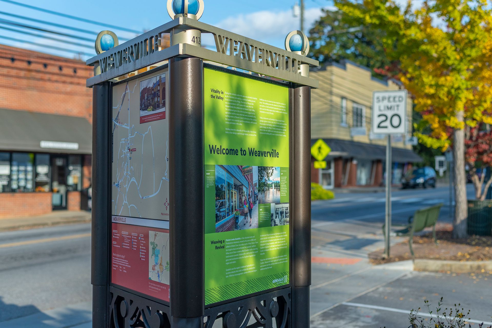 Street-side information kiosk with green and brown panels, speed limit sign in the background, sunny day.