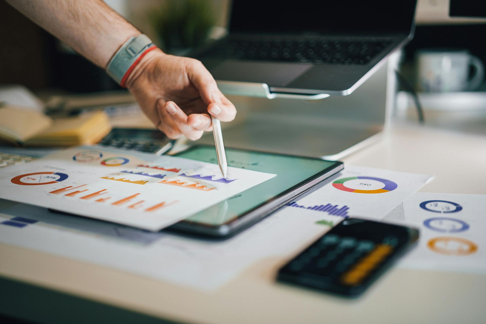 Person pointing at charts on a tablet with a laptop and calculator on a desk.