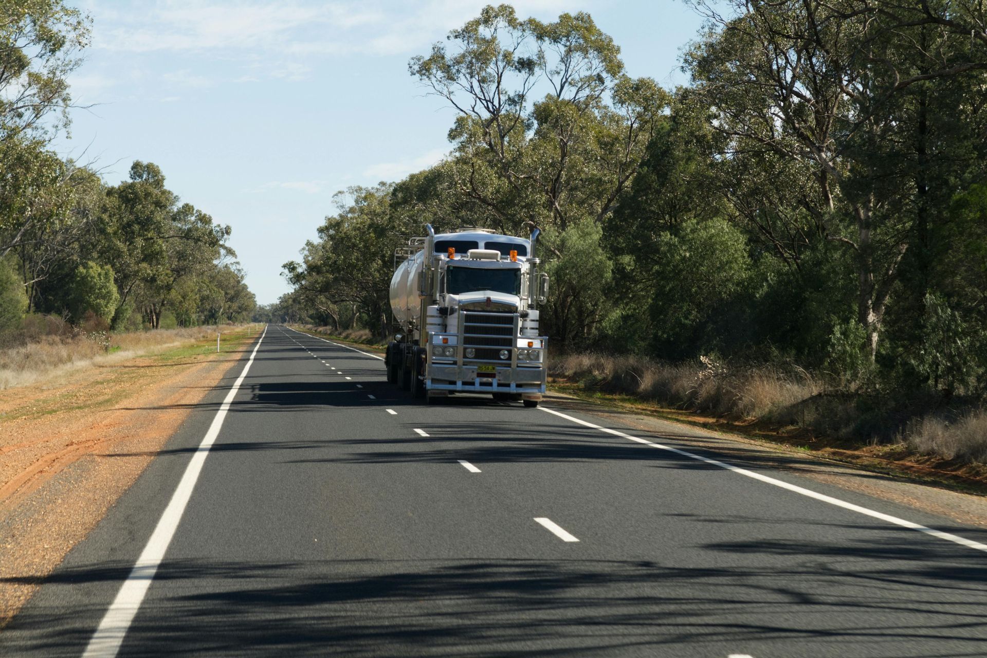 Truck traveling down a long, straight road bordered by trees under a blue sky.