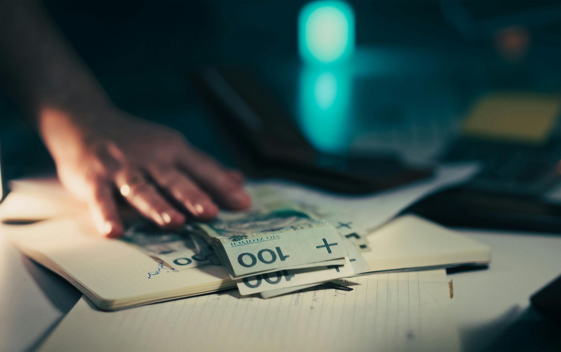 Hand reaching for a stack of Polish 100 złoty banknotes on a notebook, dimly lit.
