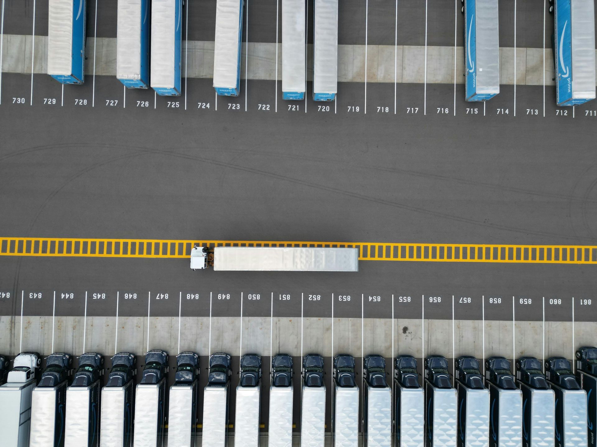 Aerial view of a truck depot with rows of parked semi-trucks; one truck is moving along a yellow line.
