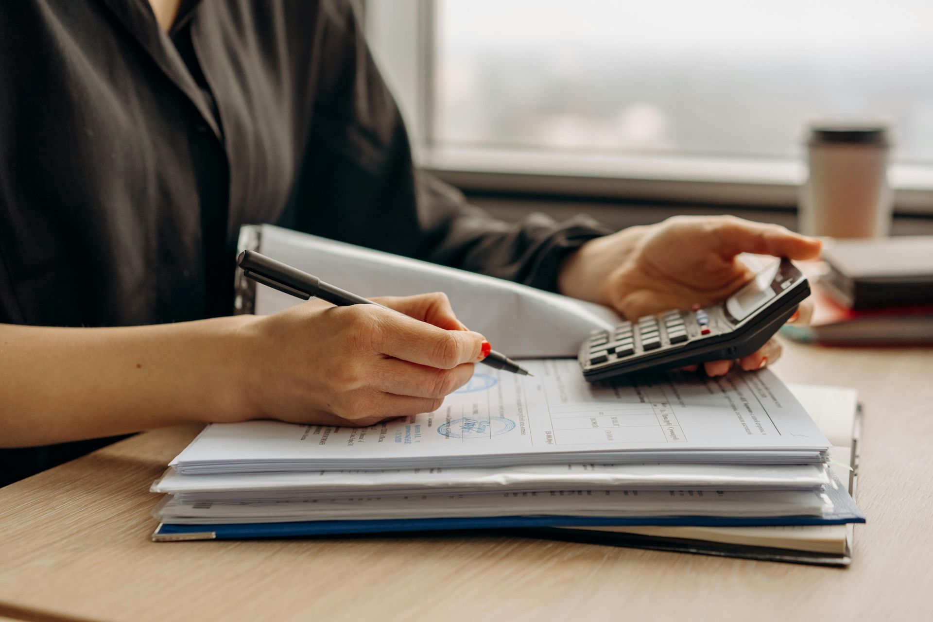 A person in a black shirt uses a calculator while holding a pen over a stack of paperwork on a desk.