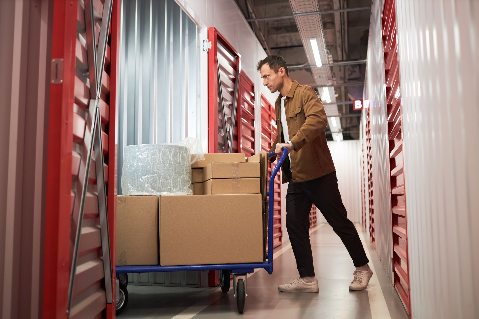 Man pushing a cart of boxes into a storage unit. Red and white hallway, neutral tones.
