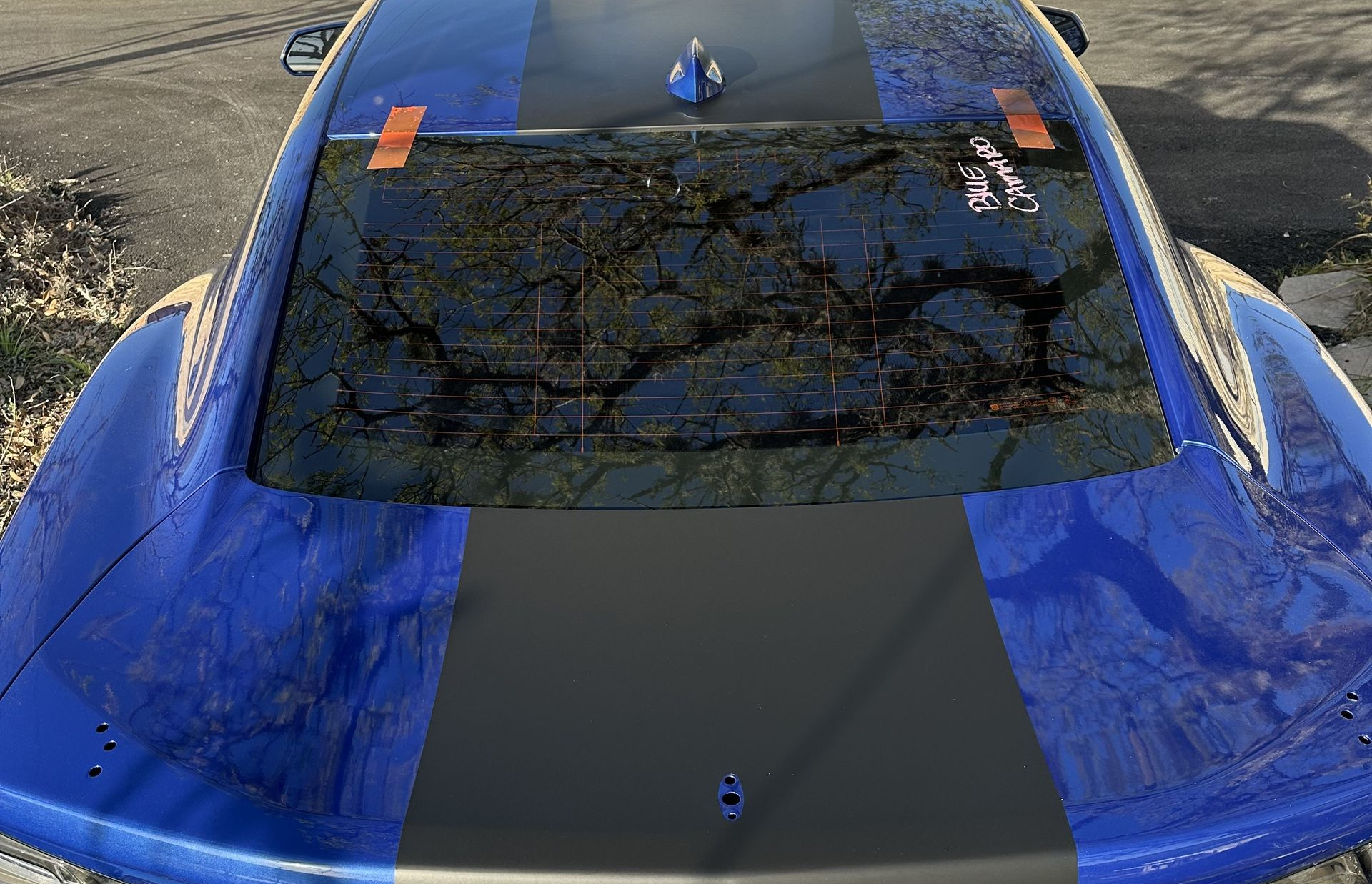 Blue car's back window shattered and taped. Black stripe on the roof. Outdoors, daylight.