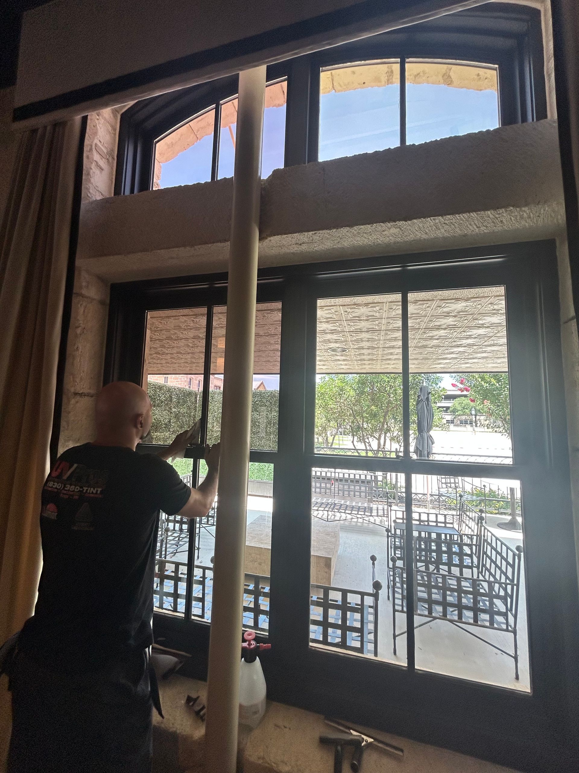 Person cleaning a window with a long pole, seen from inside the building. Tall windows overlook an outdoor patio with tables and chairs.