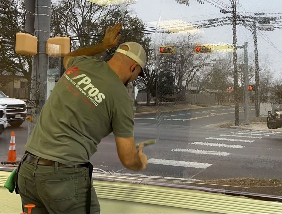 Man in a green shirt and hat squeegees a window, reflecting a street scene with traffic lights and a crosswalk.