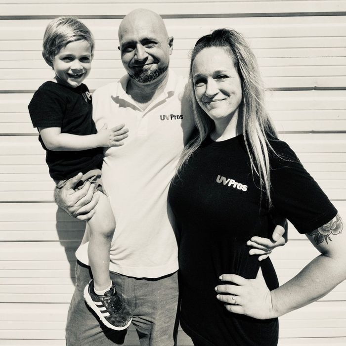 A family of three stands outside. A man holds a young boy, while a woman stands beside them. They all wear matching shirts with a logo.