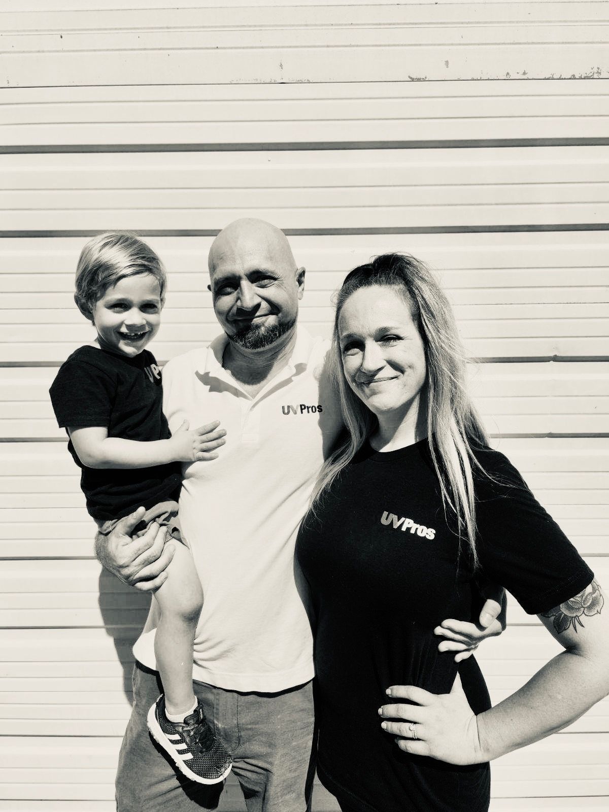 A family of three smiling in front of a white wall. Father holds a young child, mother stands beside them.