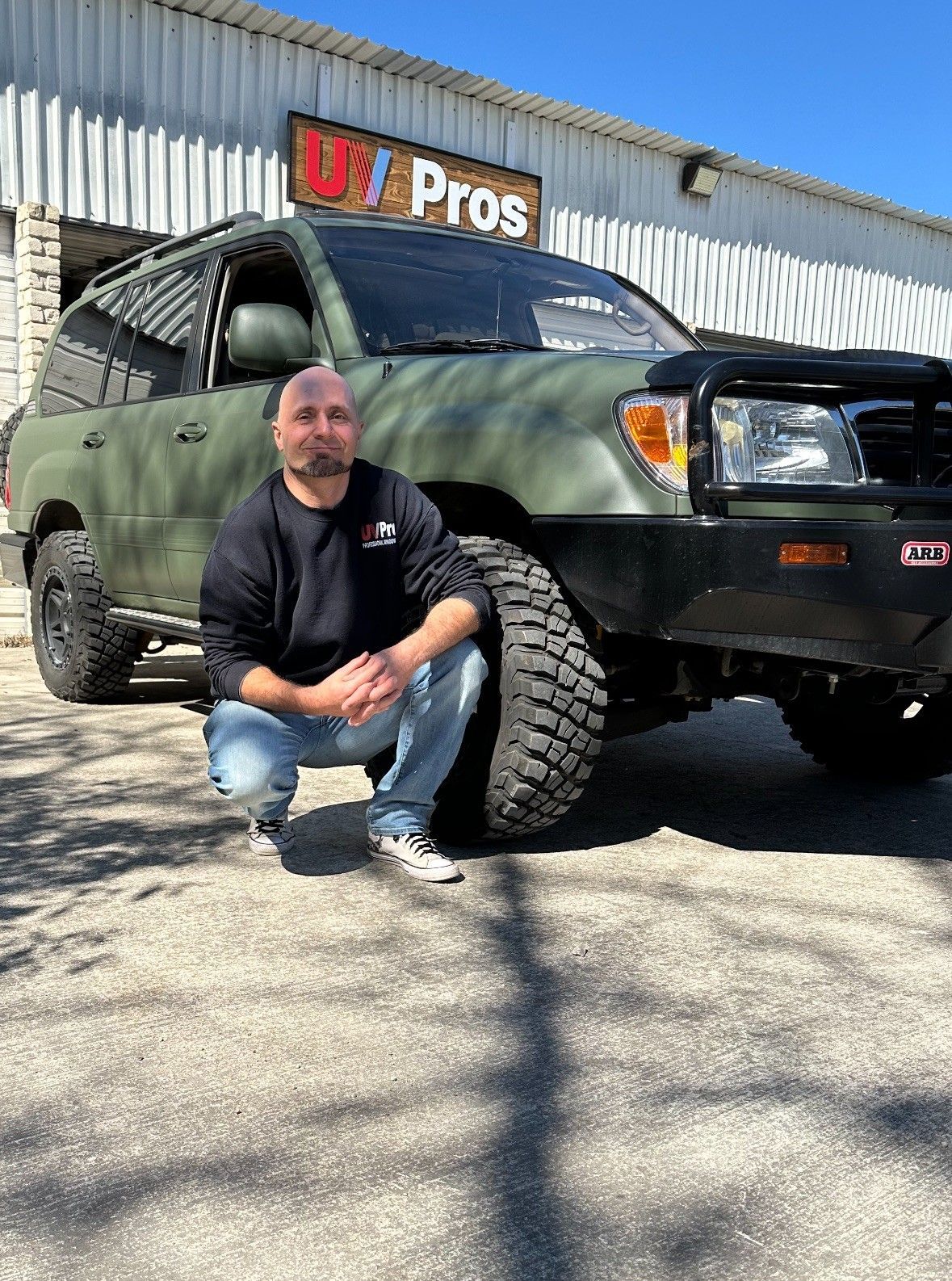 Man kneels beside a green, modified SUV with large tires. The man wears a black shirt and jeans in front of a building with 