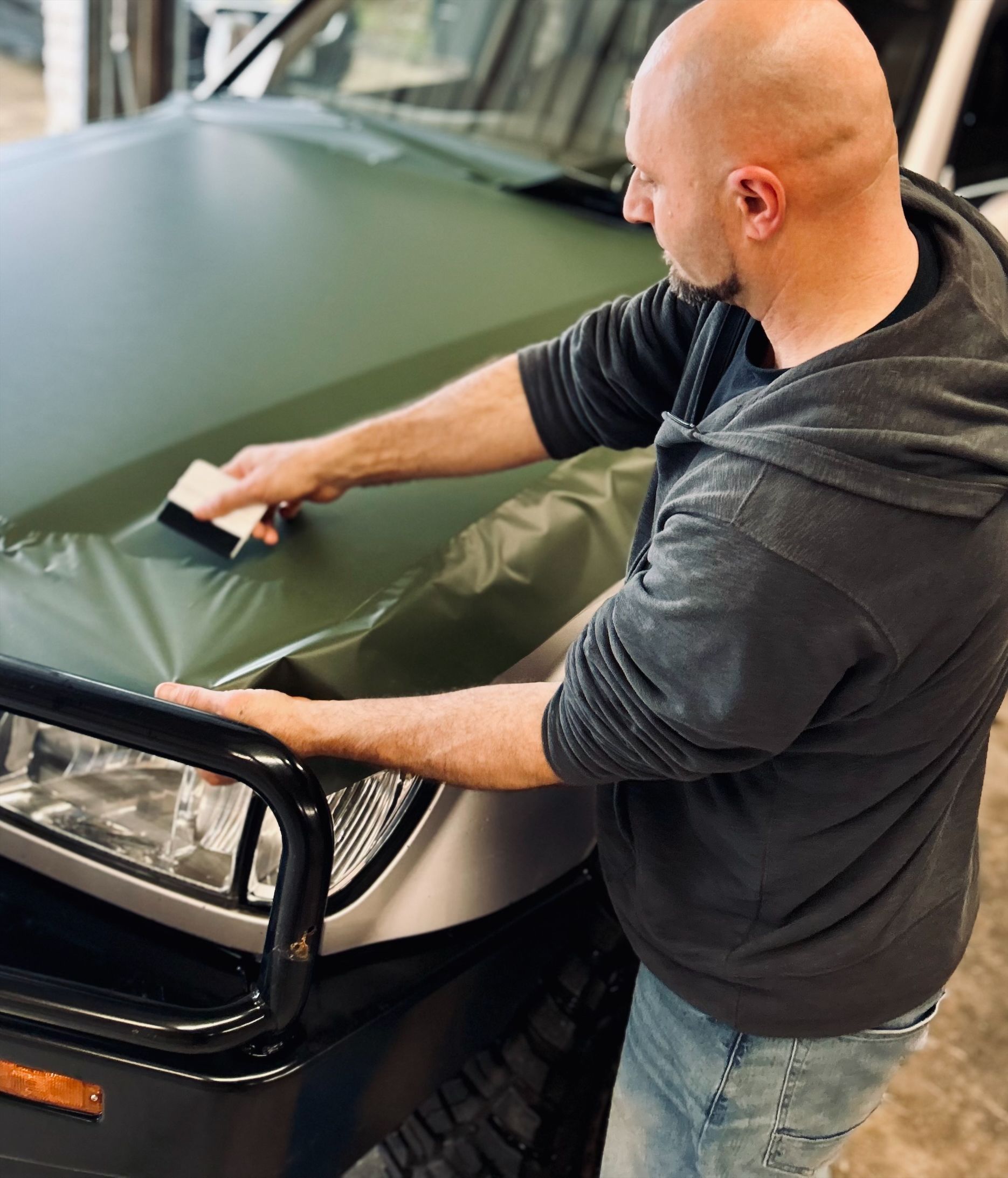 A man wrapping the hood of a green car with film, using a squeegee. He's in a garage setting.