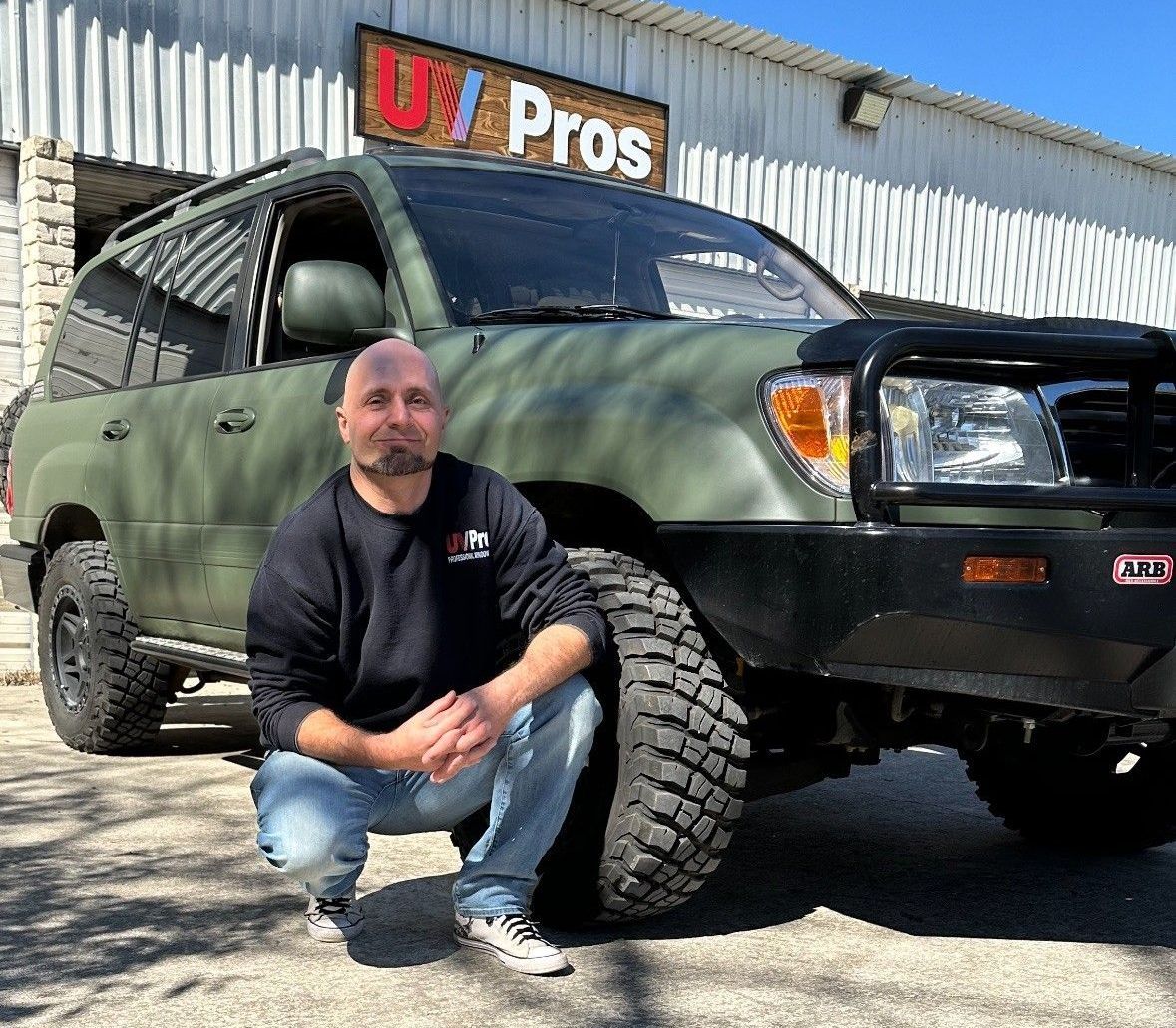 Man in front of a green SUV at UZ Pros. He is kneeling and smiling. The vehicle has black accessories and large tires.