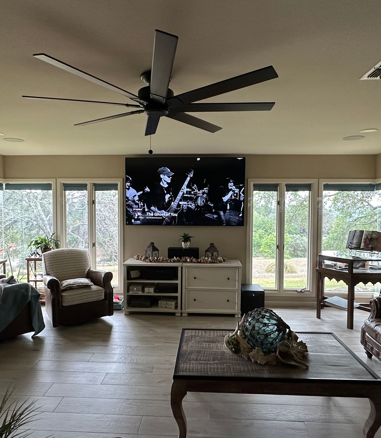 Living room with mounted TV, ceiling fan, windows, and furniture, beige walls.