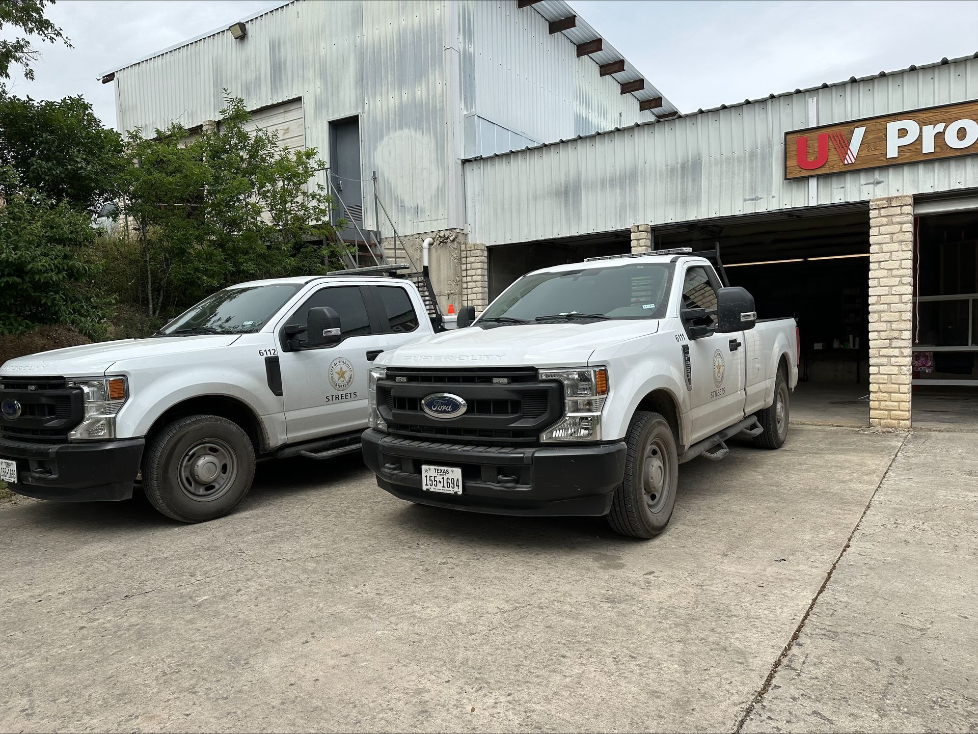 Two white Ford pickup trucks parked in front of a weathered building. One truck has a roof rack.