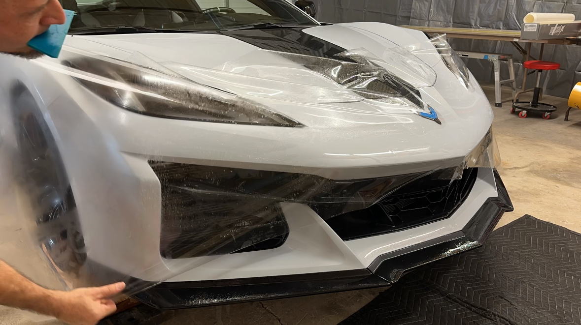 A person applying protective film to the front of a white sports car. The car is in a garage or workshop setting.