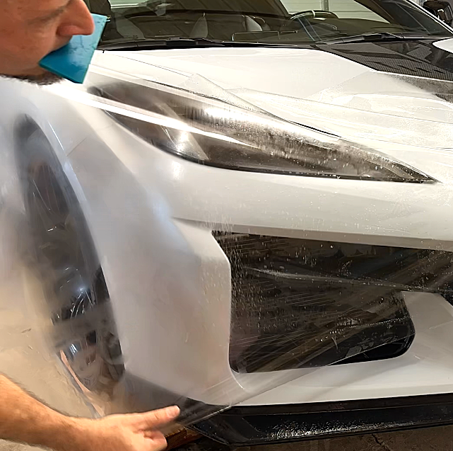 Man applying clear protective film to the front of a white sports car.