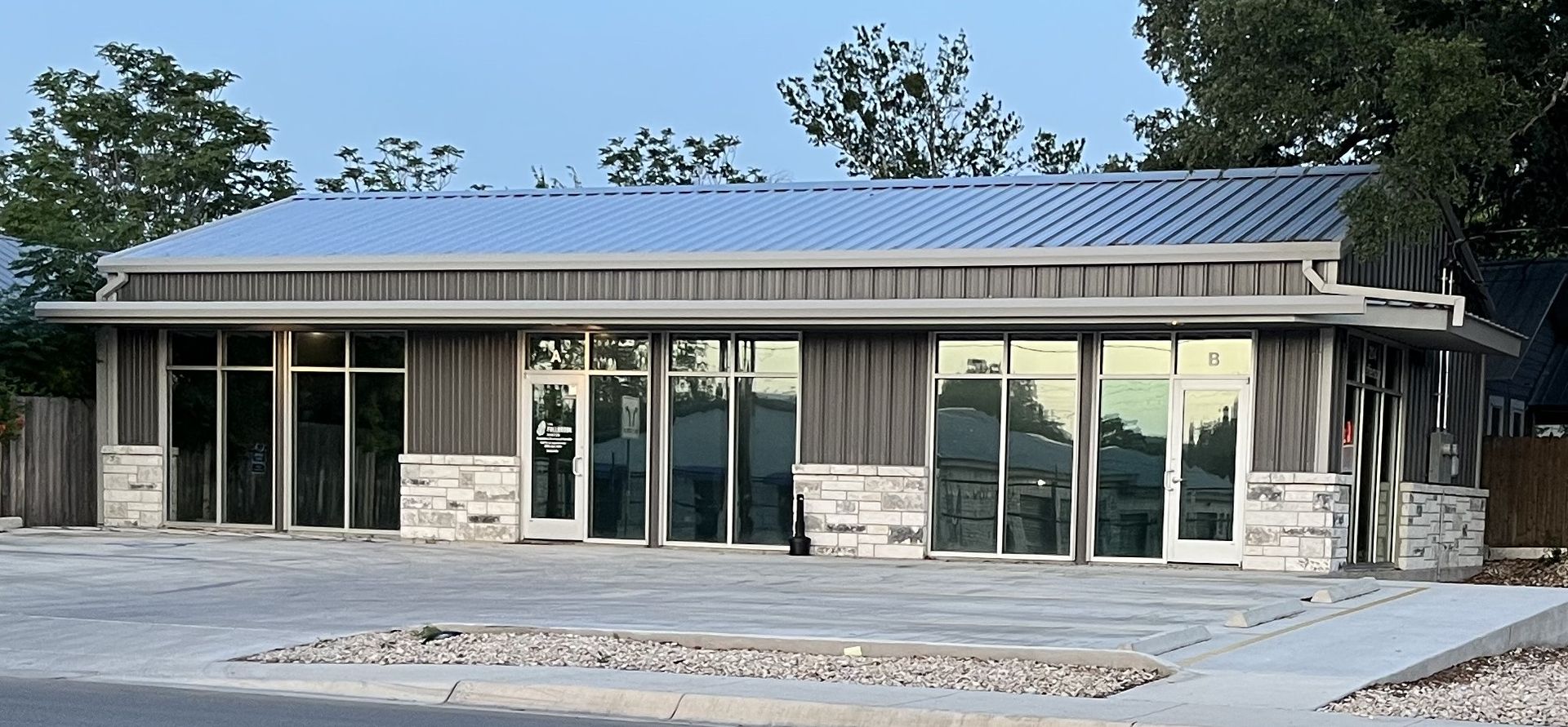 Modern building with large glass windows, gray siding, and a silver metal roof. It has light-colored stone accents.