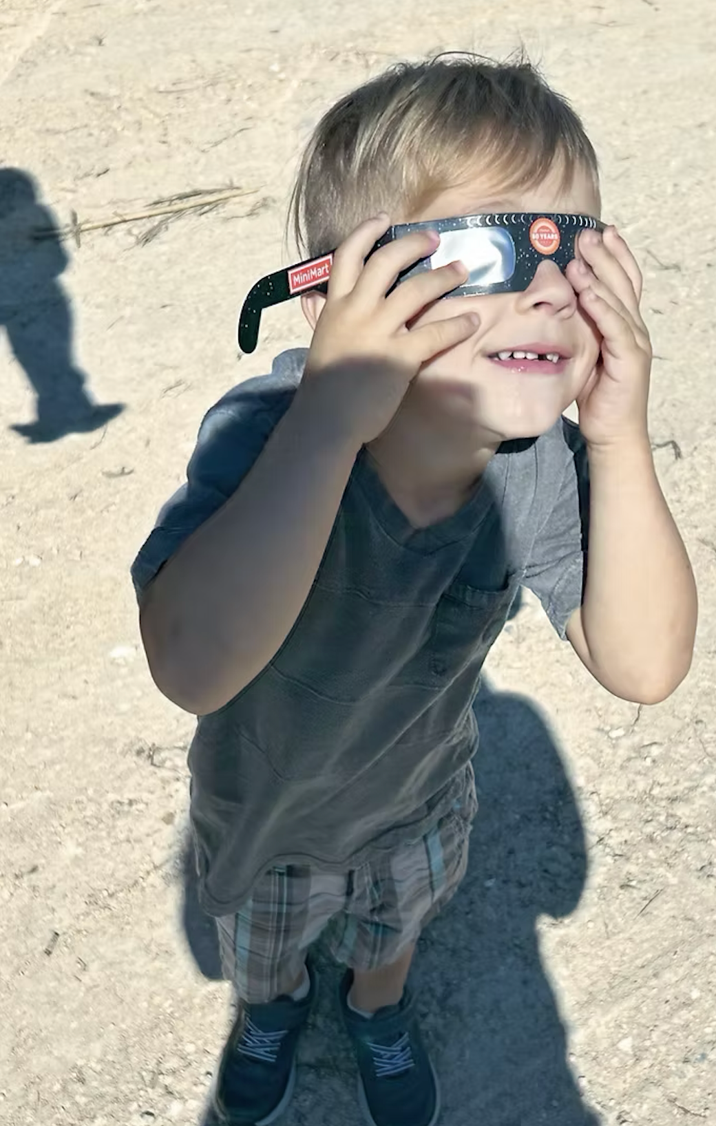 Boy wearing eclipse glasses, hands over eyes, looking up, outdoors.