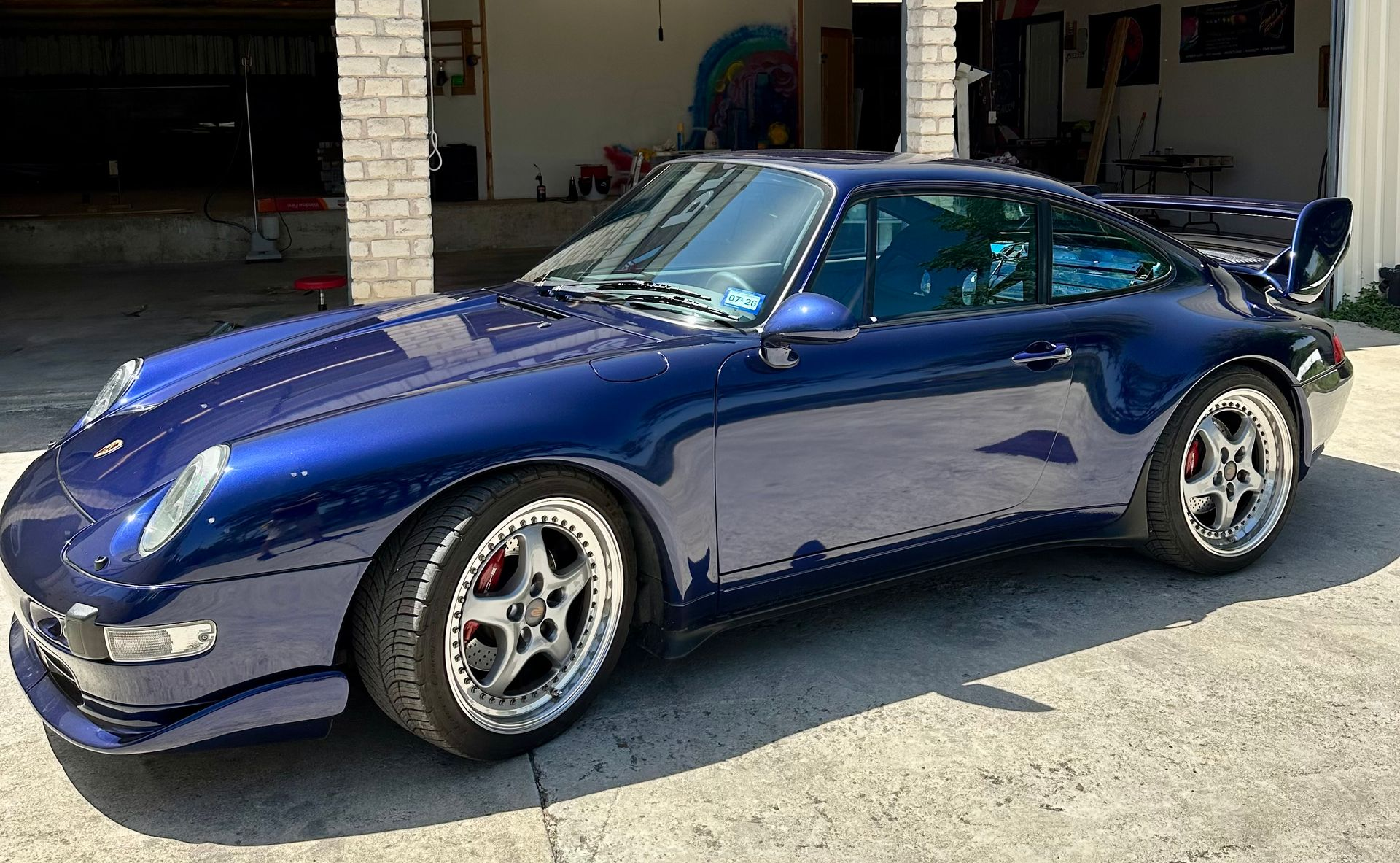 Blue Porsche 911 parked in front of a garage with chrome wheels and a rear spoiler.