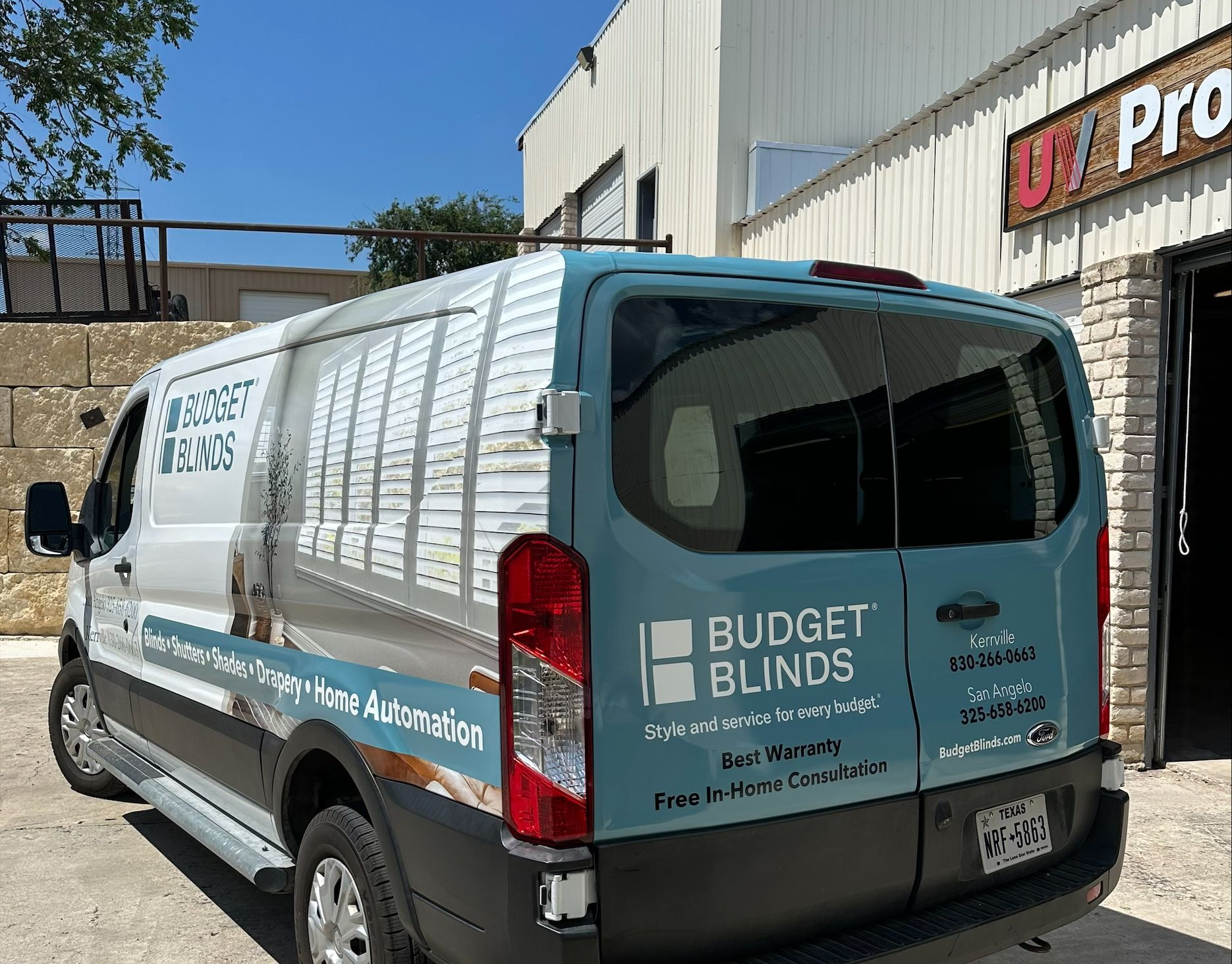 A white Budget Blinds van parked on a road, with UV Pro sign visible in the background. The van has advertising graphics for blinds and shades.