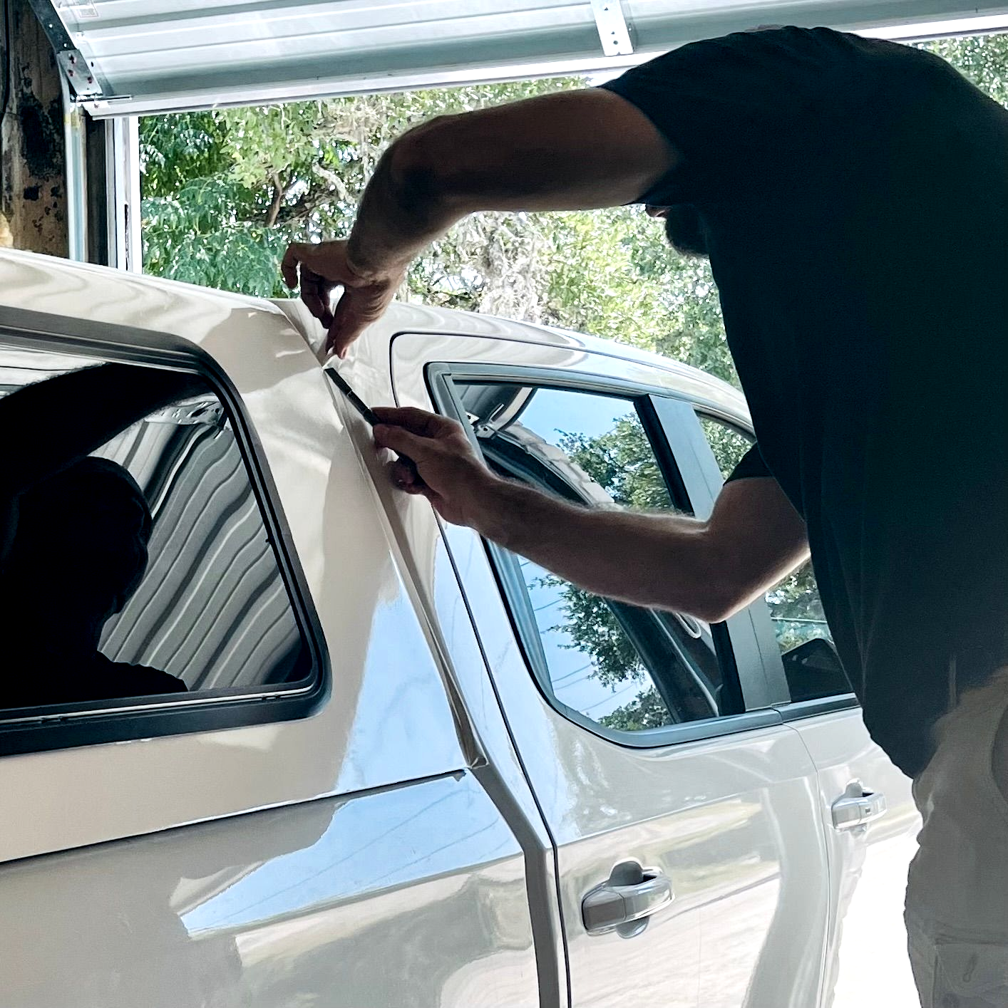 A person wearing gloves applies white vinyl wrap to the hood of a gray car in a workshop.