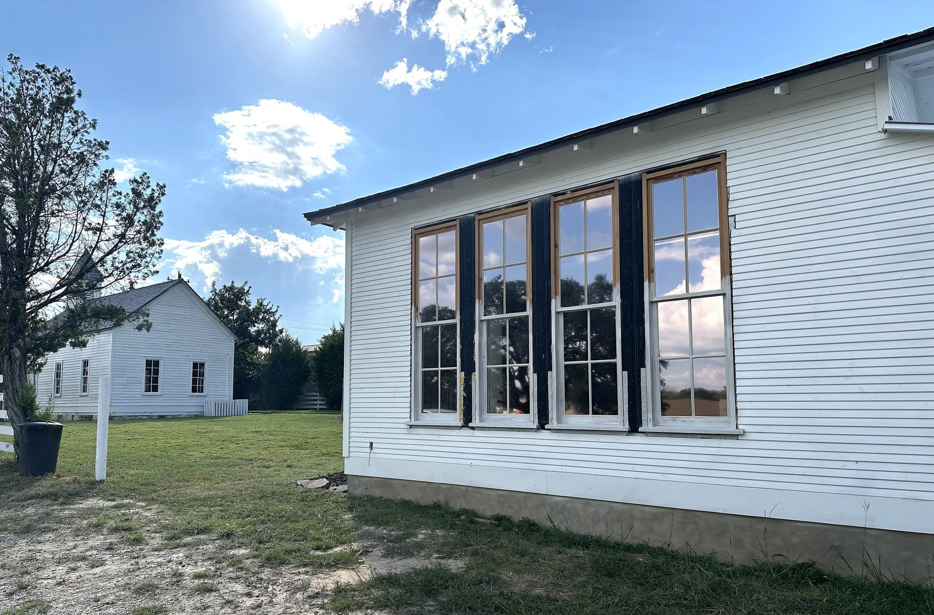 White clapboard buildings under a blue sky. One has three tall windows with dark shutters. A tree stands to the left.