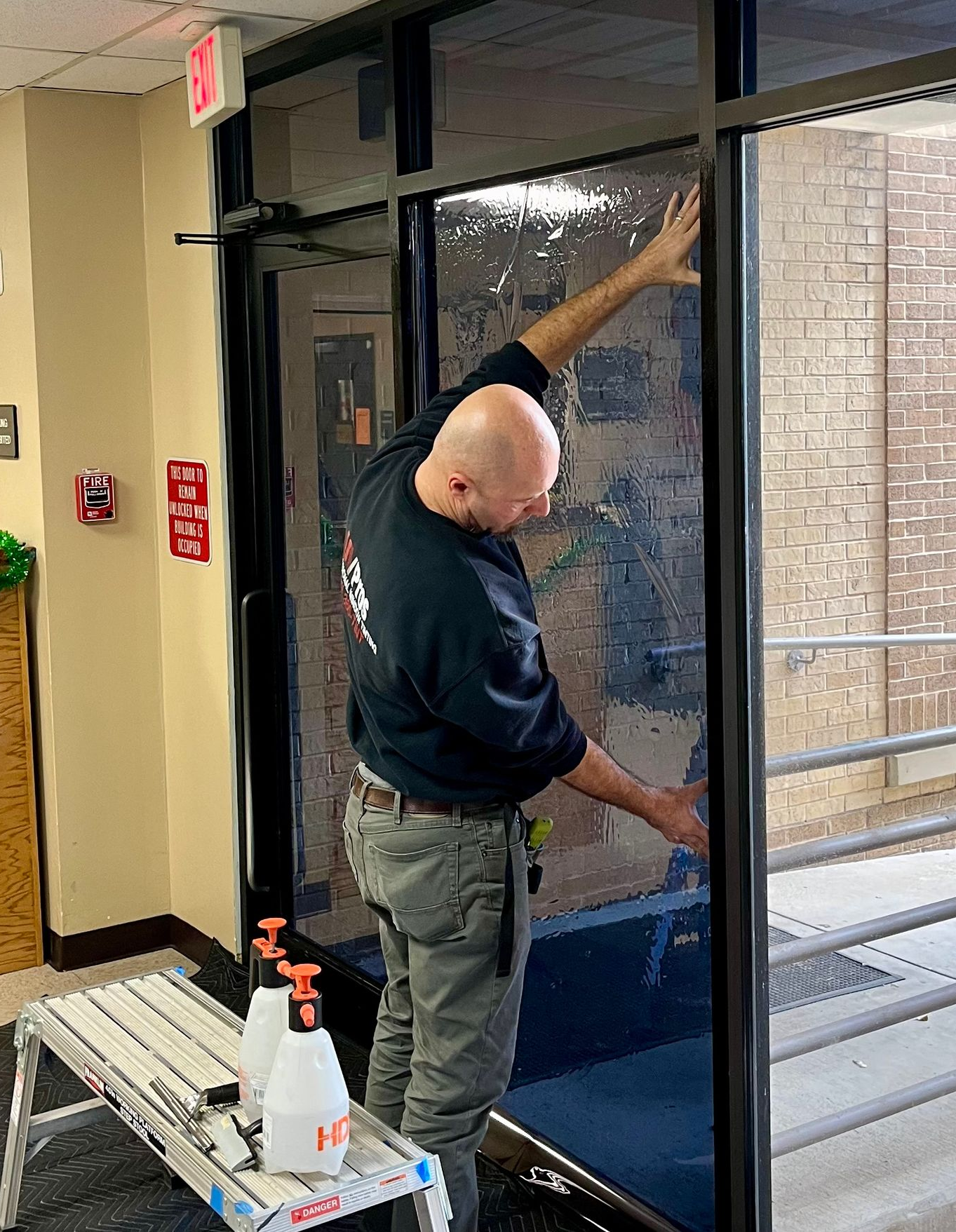 A man applying window tint to a glass door in a building hallway, spraying with a bottle and smoothing the tint.