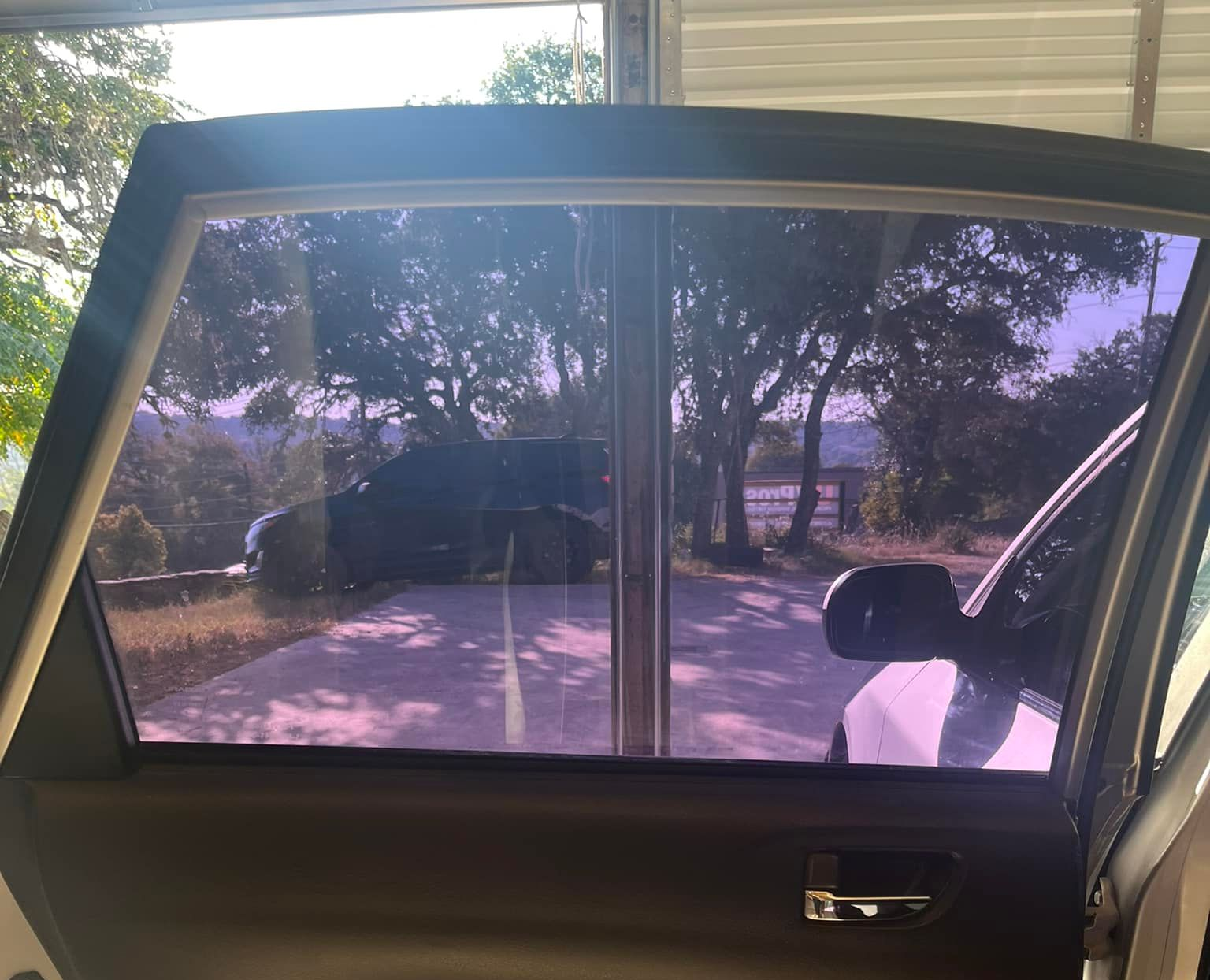 Open car door with tinted purple windows, driveway, trees, and a dark-colored car in the background.
