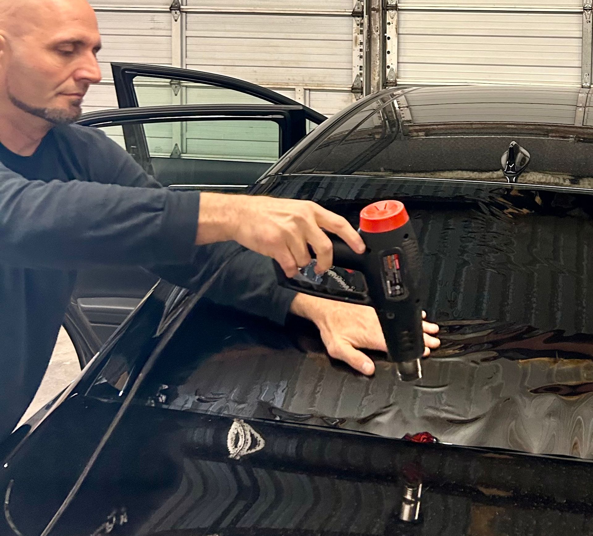 Man using a heat gun to apply tint film to a car window. Black car in a garage.
