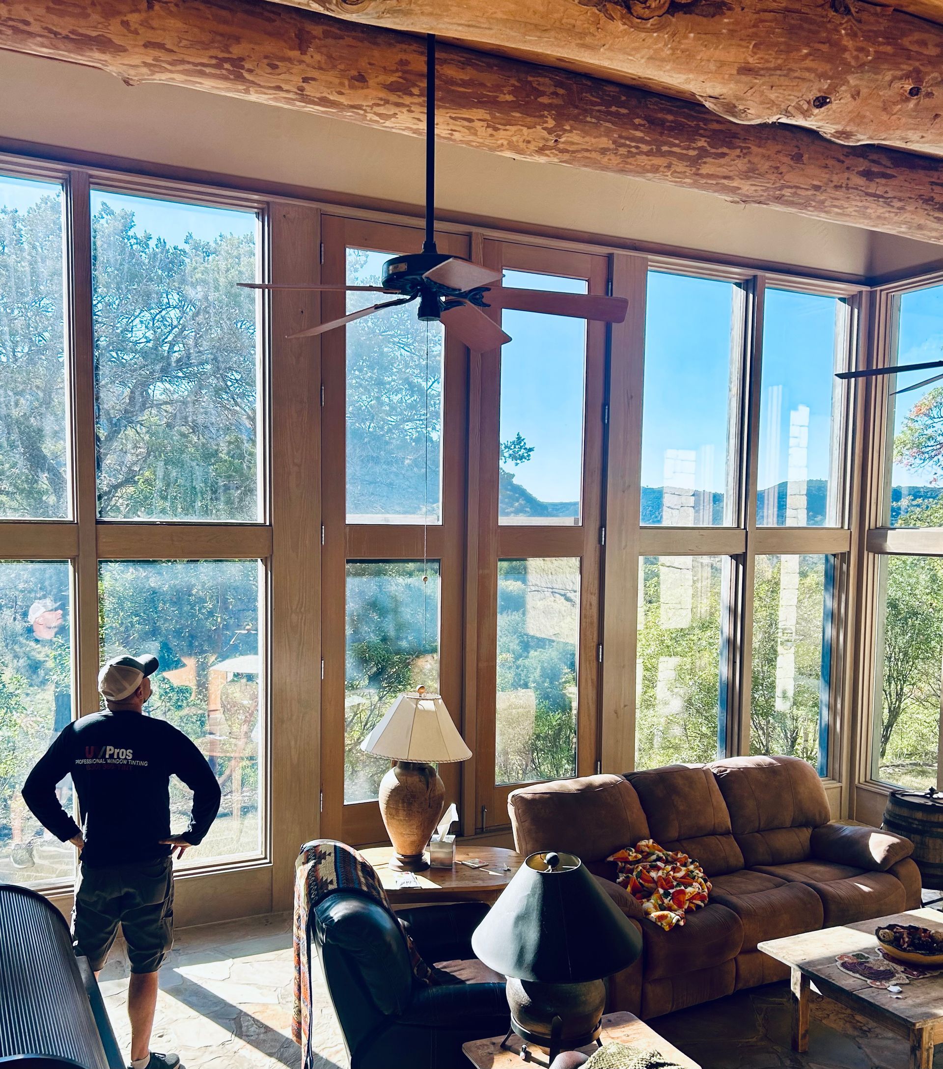 A person in a dark shirt and hat looks out large windows into a bright landscape. The windows frame a living room with rustic furniture and exposed beams.