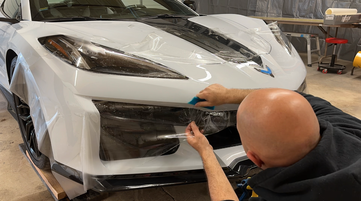 A person applying clear protective film to the front of a white Corvette in a garage.