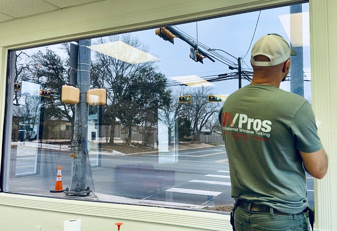 A man in a green shirt and cap looks out a large window, reflecting a street scene, light blue sky.