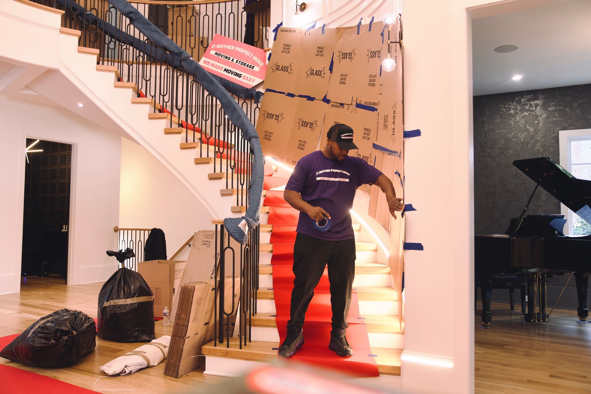 Man with black cap and purple shirt adjusts cardboard on a staircase with red carpet. A grand piano sits nearby.