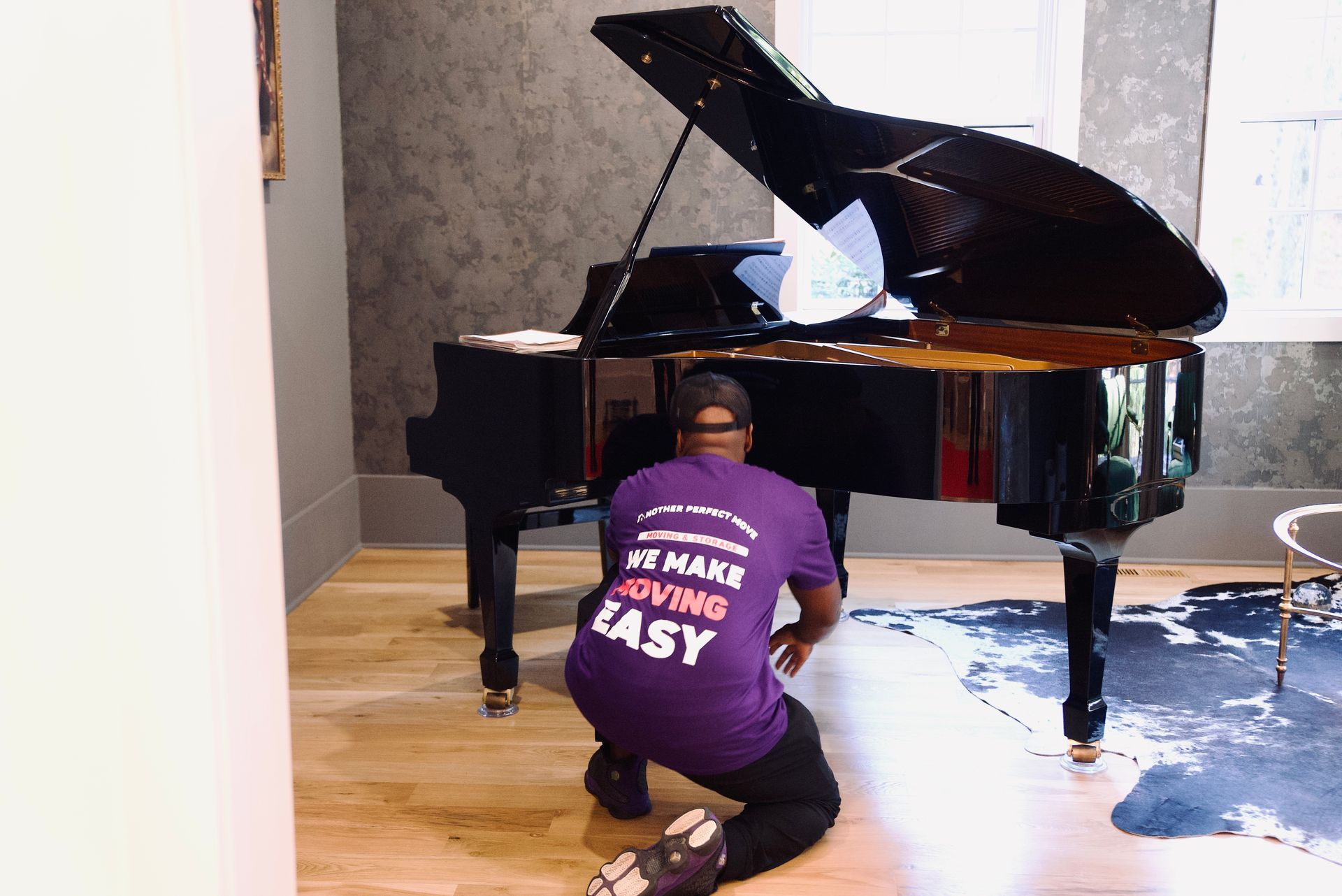 Man in purple shirt with piano, kneeling and working on the instrument in a room.