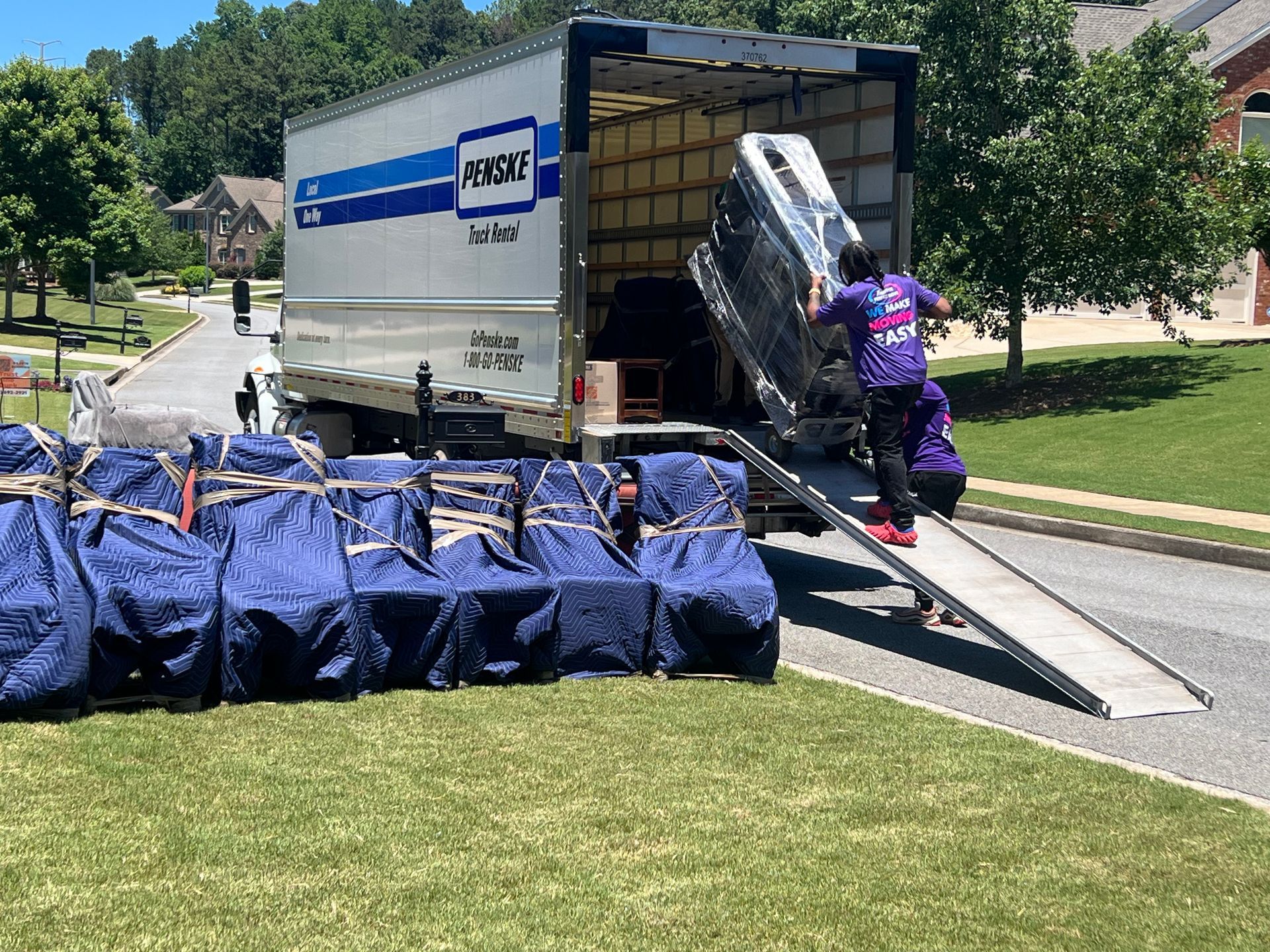 Movers unloading furniture from a truck onto a ramp on a sunny residential street.