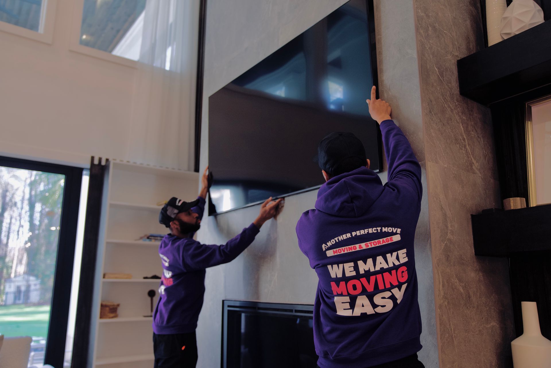 Two people in purple hoodies mount a large TV on a living room wall.