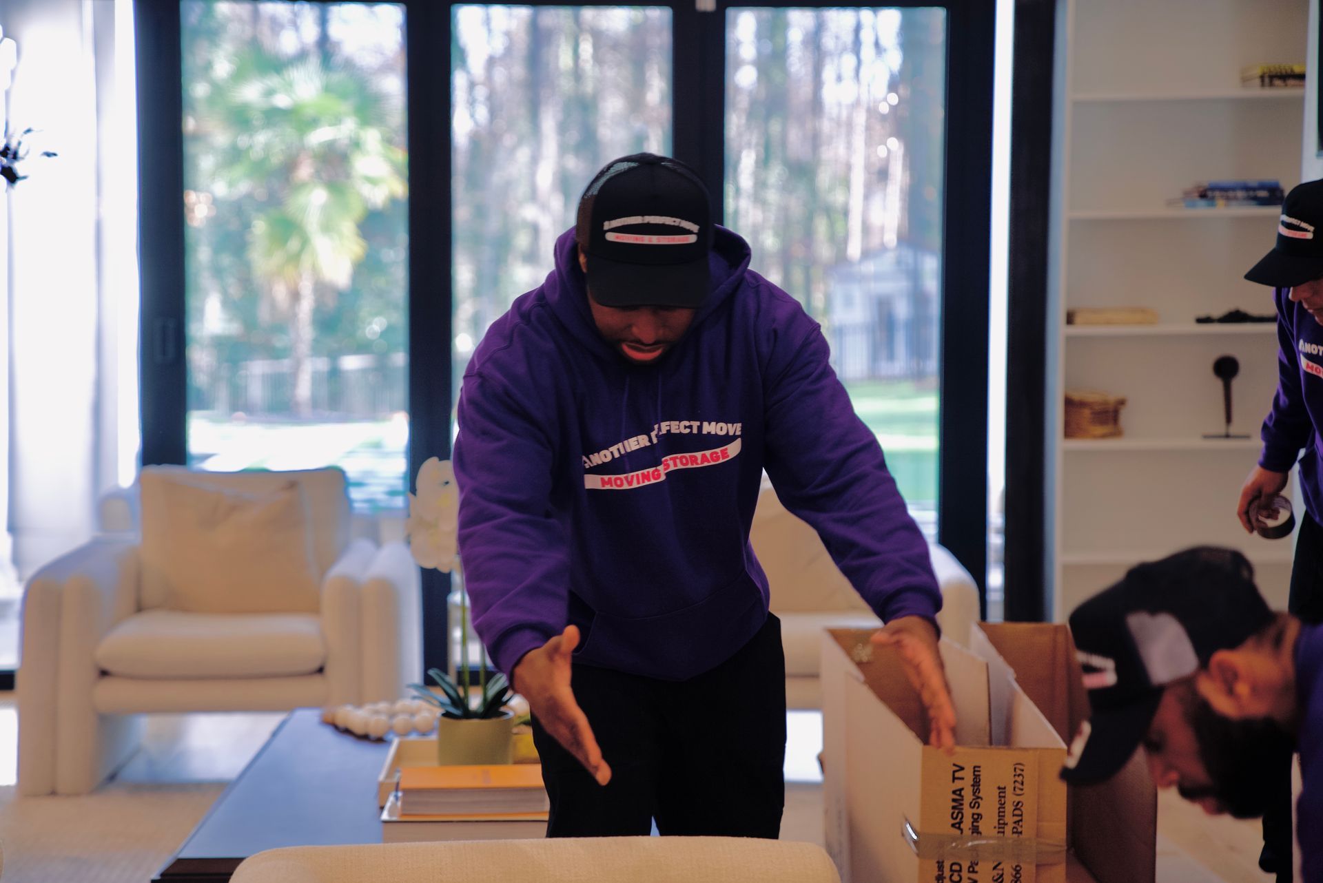 A man in a purple hoodie is holding a cardboard box in a living room.