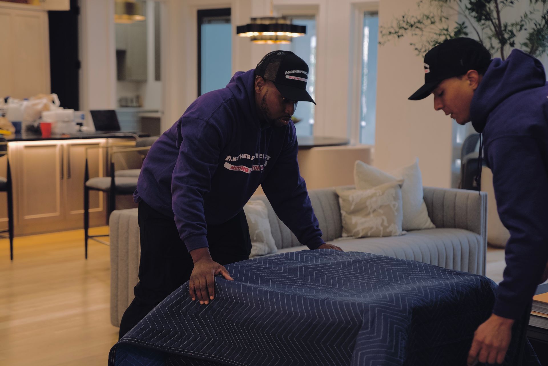 Two men in purple hoodies move a wrapped upholstered ottoman in a modern living room.