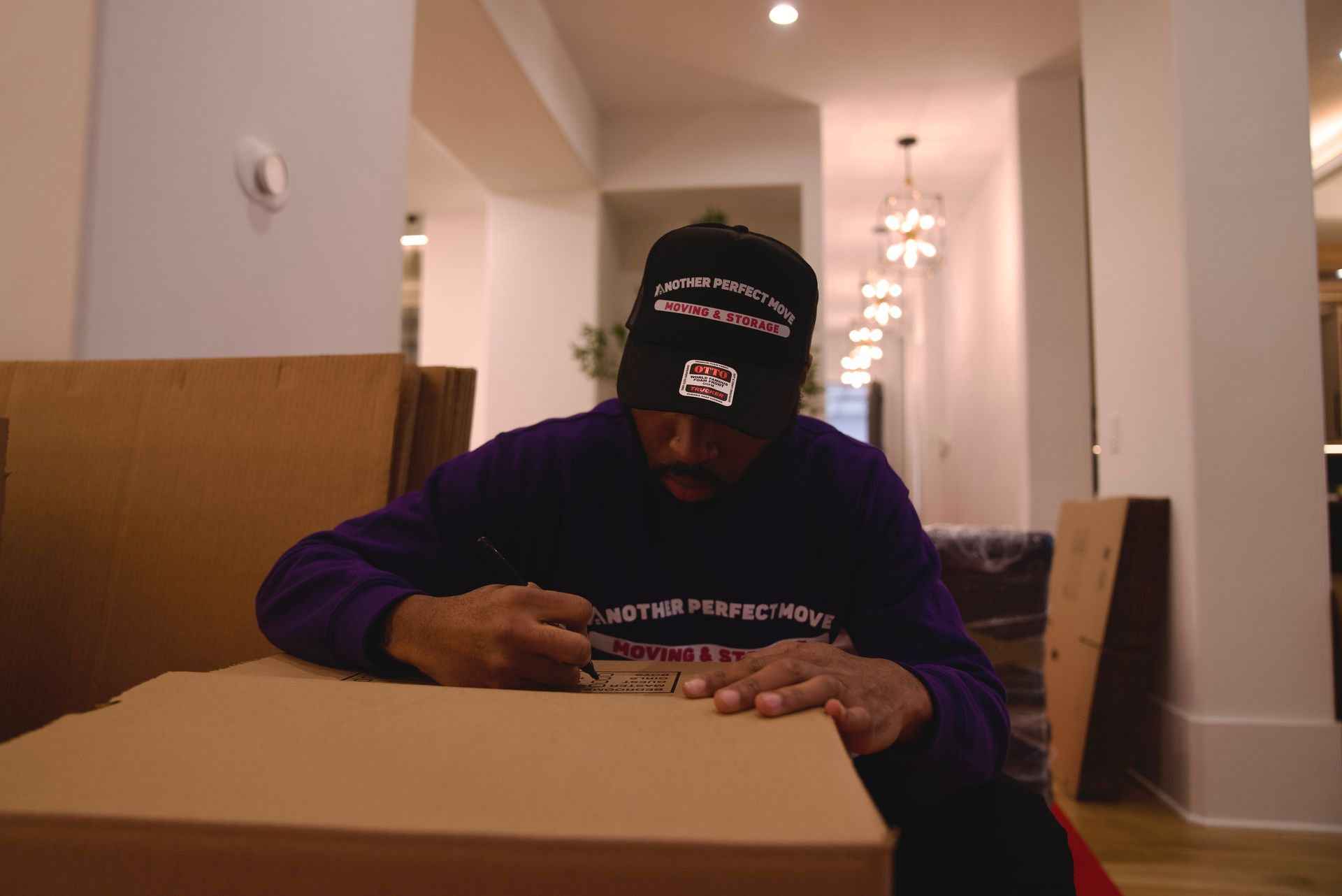 A man wearing a purple shirt and a black hat is opening a cardboard box.