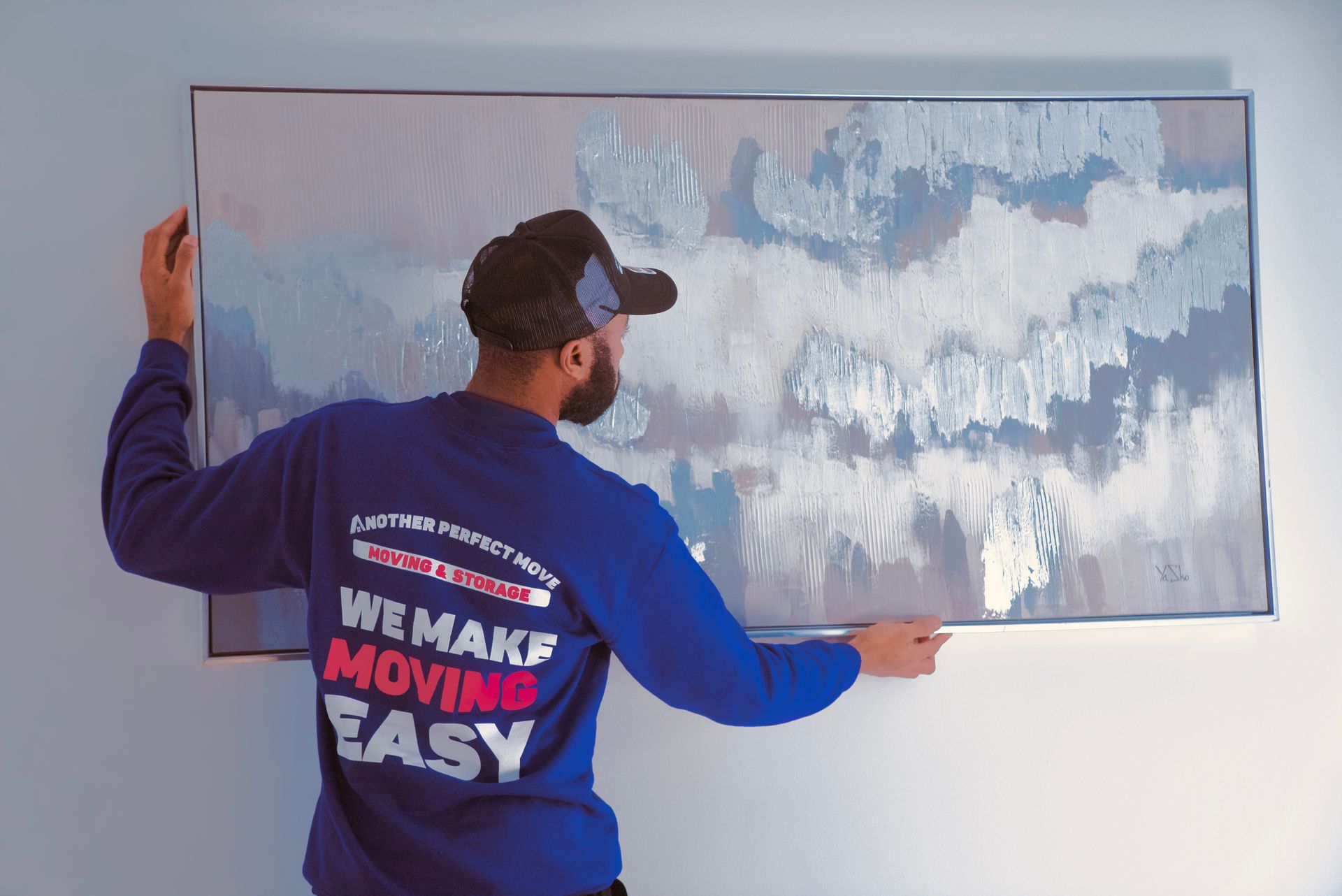 A man in a blue shirt is hanging a painting on a wall.