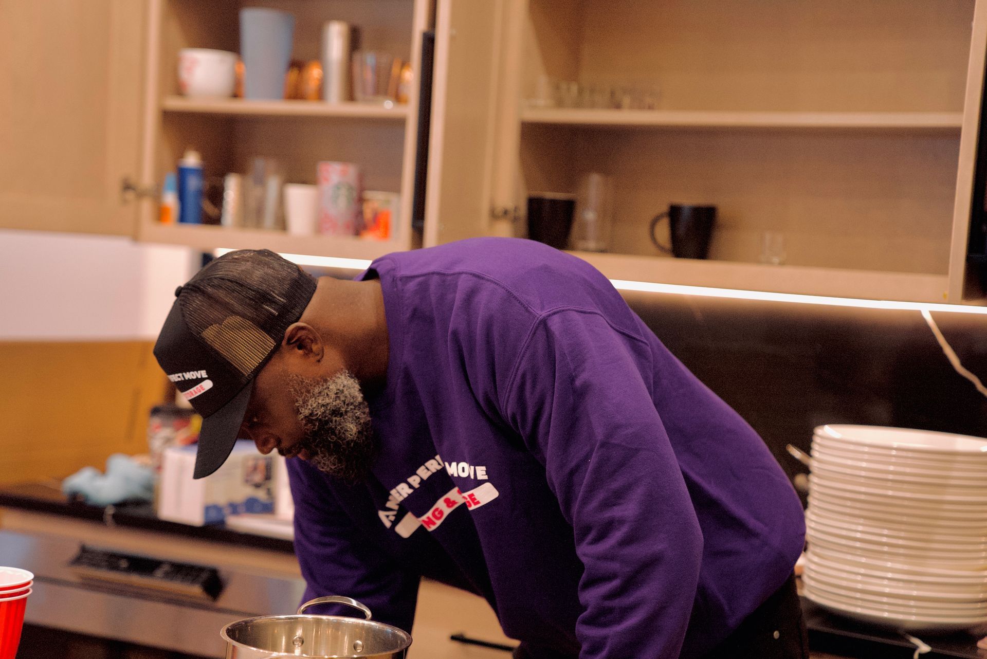 A man in a purple shirt and hat is cooking in a kitchen.