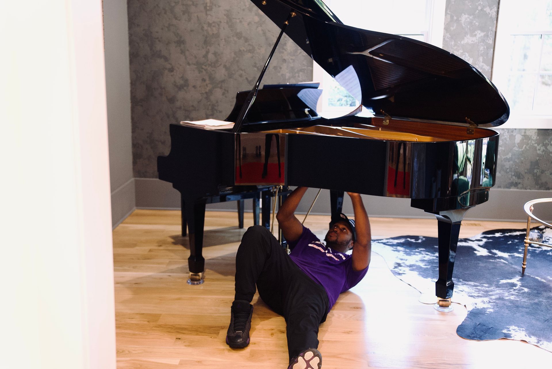 A man in a purple shirt is laying on the floor under a piano.