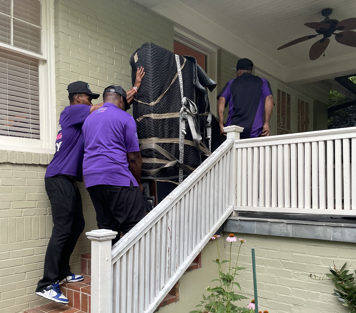 A group of men in purple shirts are carrying a piano up stairs