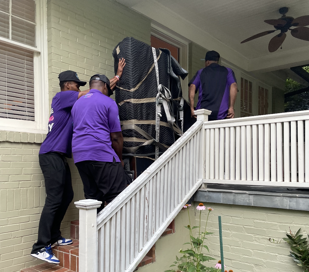 A group of men in purple shirts are carrying a piano up a set of stairs