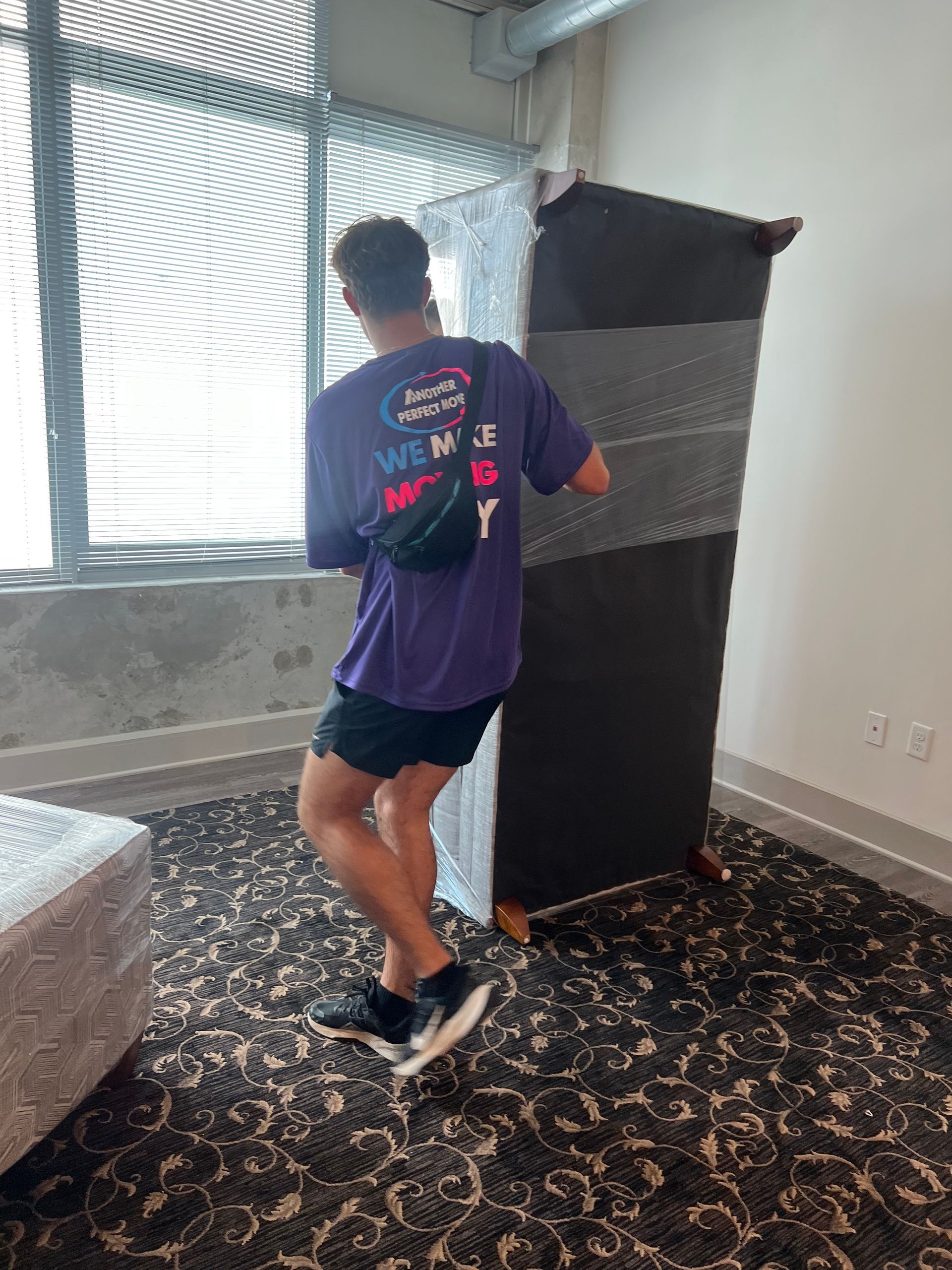 A man in a purple shirt is carrying a piece of furniture in a room.