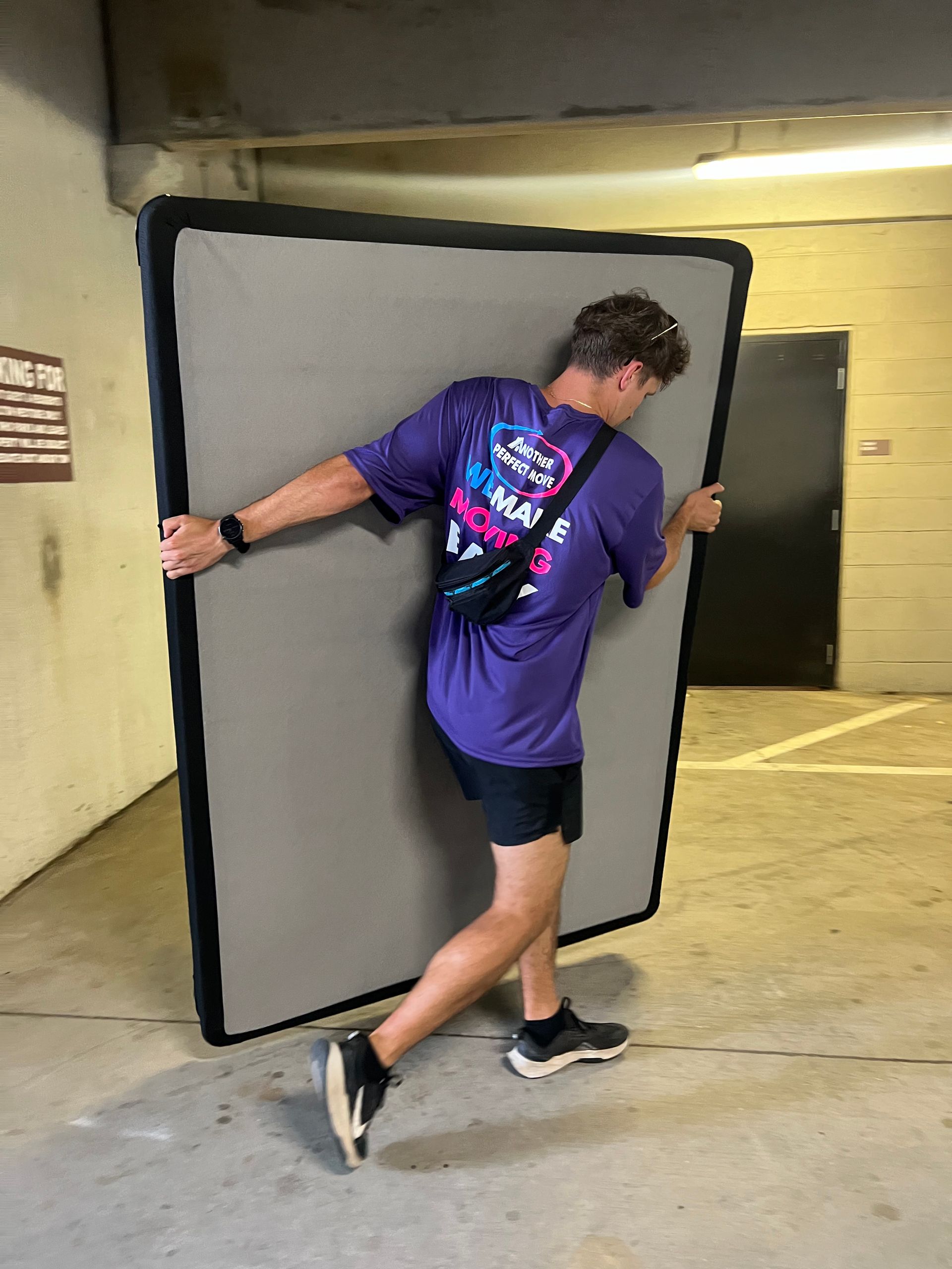 A man in a purple shirt is carrying a mattress in a parking garage.